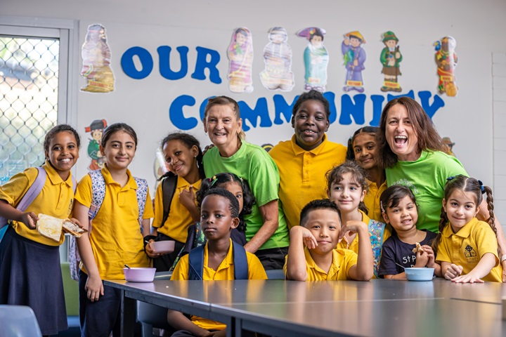 Group of children and adults gathered around a table in a classroom under a wall display reading “Our Community.”