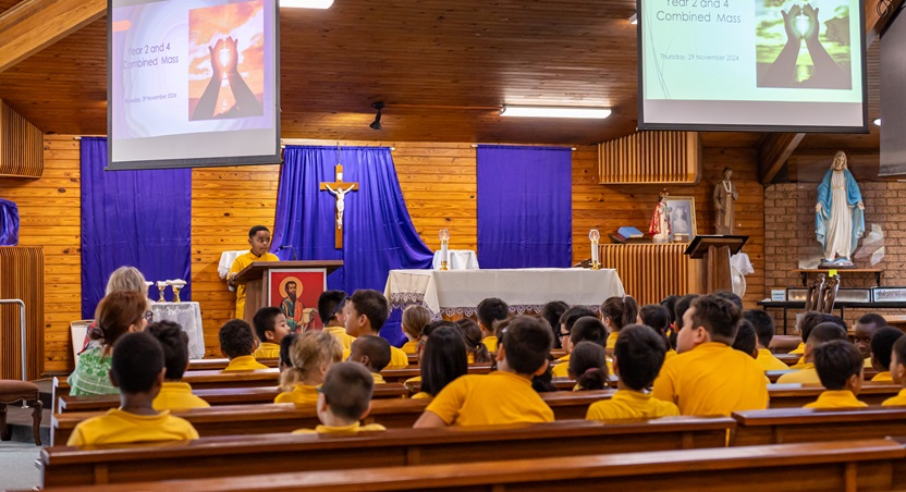 Students seated in a church facing the altar during a liturgy, with a student speaking at a lectern and projected slides displayed at the front.