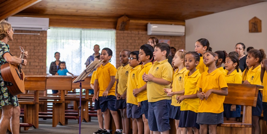 Students in yellow uniforms singing as a choir inside a church while an adult plays guitar at the front, with others seated in the pews behind them.