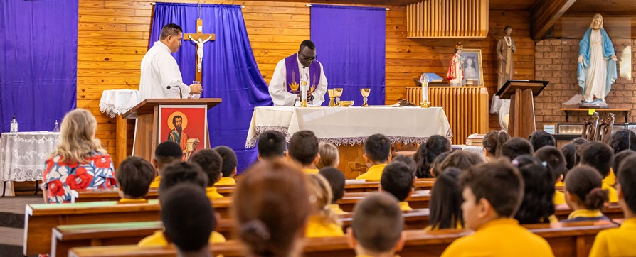 Students seated in a church while clergy lead a liturgy at the altar, with religious symbols and purple cloths displayed.