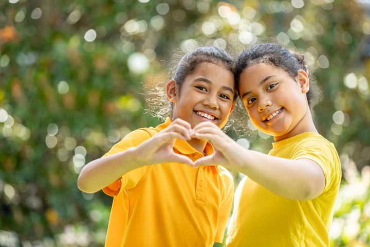 Two children in yellow shirts forming a heart shape with their hands outdoors.