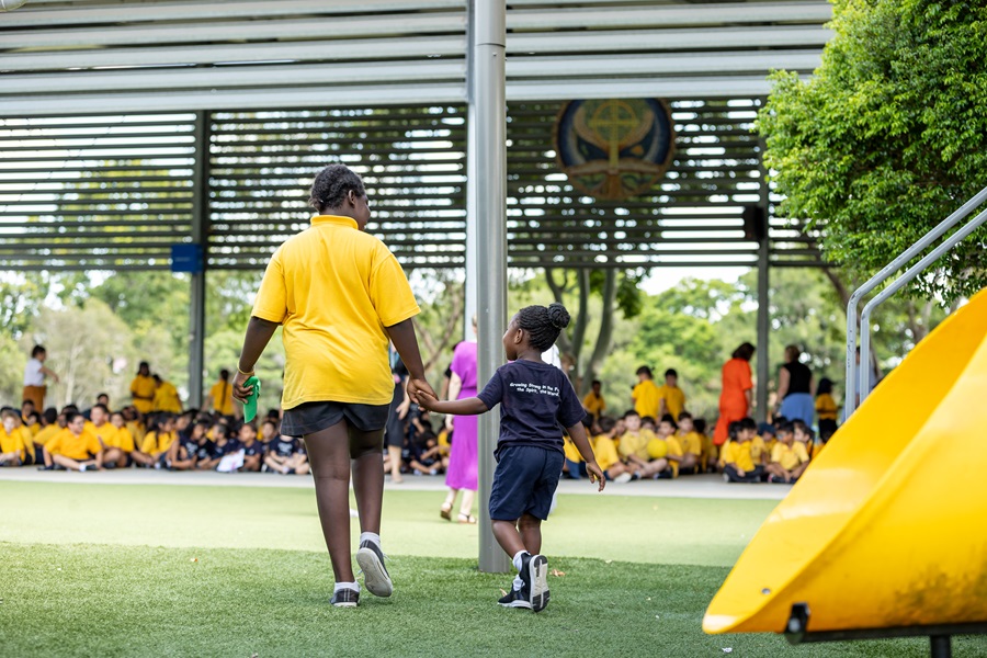Two children holding hands walking across a grassy area, with a large group of students seated under a covered space in the background.