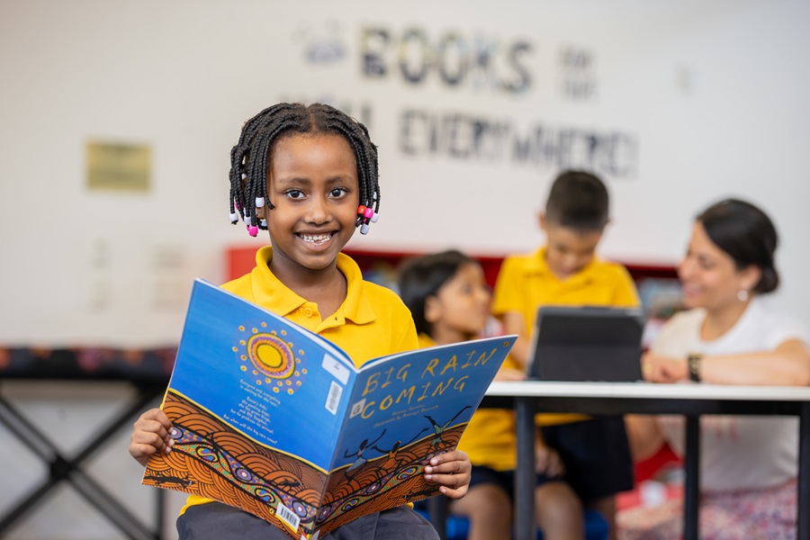 Student holding and reading a colourful picture book in a classroom, with other students and an adult using devices in the background.