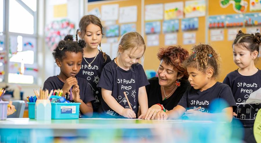 Adult assisting a group of young children at a classroom table during a hands‑on learning activity, with art materials and student work displayed on the walls behind them.