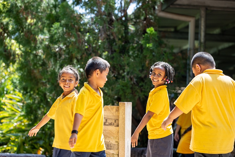 Four children in yellow school uniforms walking together outdoors in a garden area beside school buildings.
