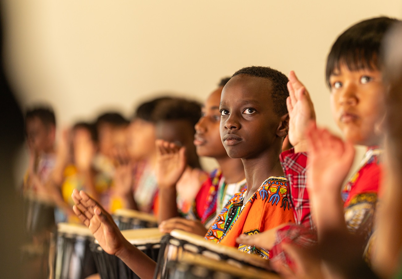 Children seated in a row playing hand drums during a music activity, wearing brightly patterned clothing.