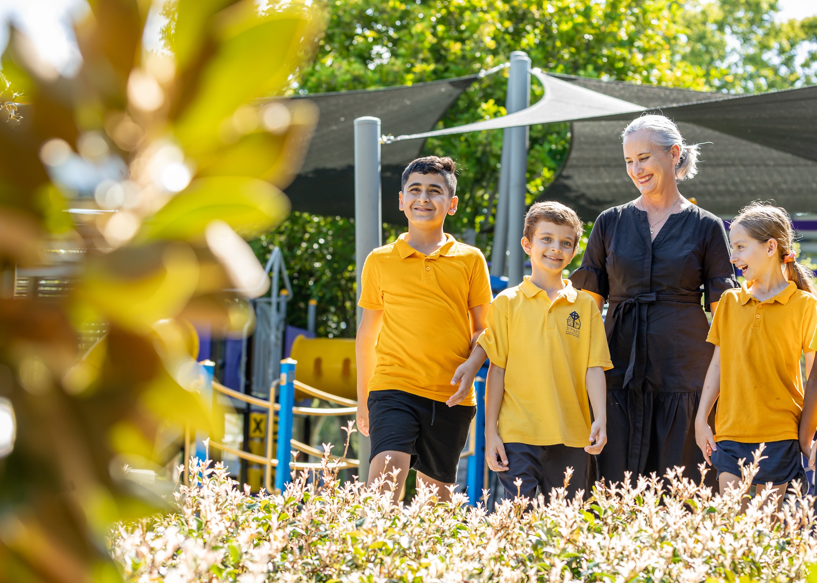 Students in yellow uniforms walking outdoors with an adult near playground equipment and garden landscaping.