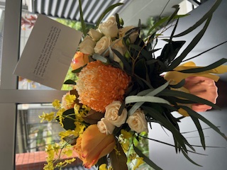 Bouquet of flowers in soft orange, yellow and white tones arranged in a vase indoors, with a card attached to the arrangement.