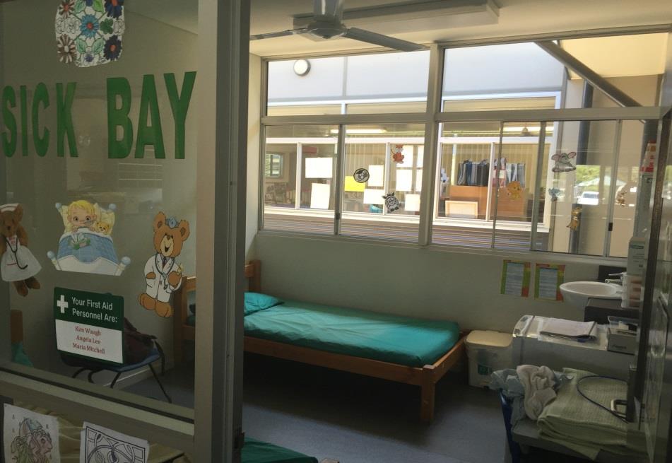 School sick bay room with a bed, sink and storage, viewed through a glass door labelled “Sick Bay” with first aid staff names listed.